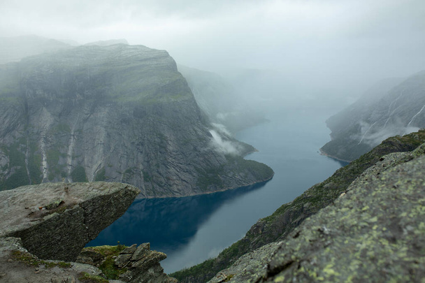man on the edge of a troll tongue in norway in rainy weather on a background of mountains and a river - Foto, imagen