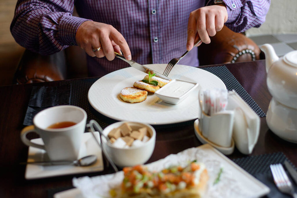 ранковий сніданок або вечеря в ресторані. стіл з напоями та їжею. чоловік руками нарізає сирники ножем і виделкою. вибірковий фокус
 - Фото, зображення