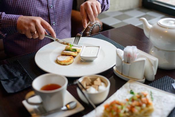 Aamiainen tai brunssi ravintolassa. pöytä juomia ja ruokaa. mies kädet leikattu juustokakkuja veitsellä ja haarukalla. valikoiva kohdentaminen
 - Valokuva, kuva