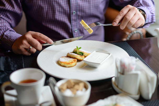 πρωινό ή brunch στο εστιατόριο. τραπέζι με ποτά και φαγητό. άνθρωπος χέρια κομμένα cheesecakes με ένα μαχαίρι και πιρούνι. επιλεκτική εστίαση - Φωτογραφία, εικόνα