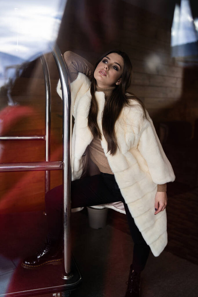 Sexy brunette girl standing near hotel luggage cart. Luggage Trolley at the hotel. Glamorous young woman wearing stylish white fur coat. Fashion, fur, hotel, travel and beautiful people concept.. - Photo, Image