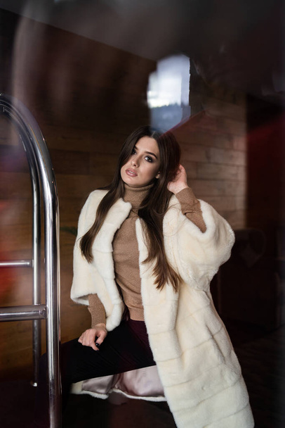 Sexy brunette girl standing near hotel luggage cart. Luggage Trolley at the hotel. Glamorous young woman wearing stylish white fur coat. Fashion, fur, hotel, travel and beautiful people concept.. - Foto, imagen