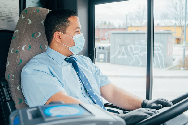 νεαρός ισπανόφωνος οδηγός λεωφορείου άνθρωπος έχει μπλε μάσκα ιατρικής προστασίας και μαύρα γάντια στα χέρια για να προστατεύσει τον εαυτό του από covid 19. οδηγός με μάσκα κοιτάζει το δρόμο. - Φωτογραφία, εικόνα