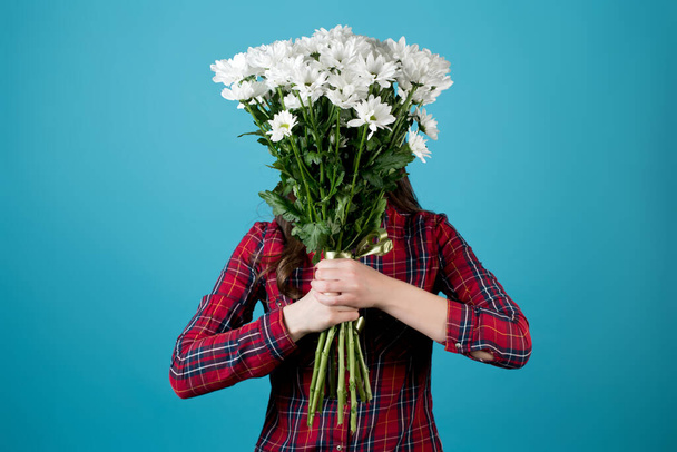 Girl in a red checkered shirt covers her face with a large bouquet of white chrysanthemums on a blue background - Photo, Image