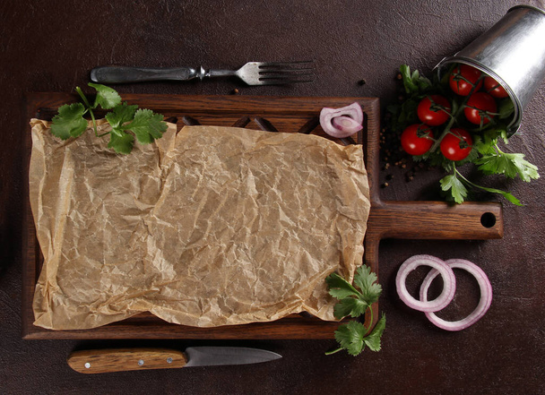 Dark wooden board with parchment on a dark brown background, a fork and knife, cherry tomatoes in a bucket and fresh herbs, coriander and parsley, onion rings and spices. Background image, copy space - Foto, Imagen
