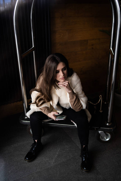 Sexy brunette girl sit on hotel luggage cart. Luggage Trolley at the hotel. Glamorous young woman wearing stylish white fur coat. Fashion, fur, hotel, travel and beautiful people concept.. - Foto, Imagen