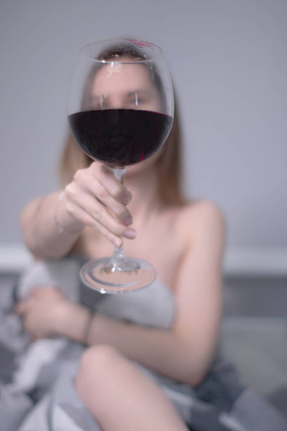 happy beautiful slender half-naked girl shows us a huge glass of red wine, in bed with gray sheets in triangles - Foto, Bild