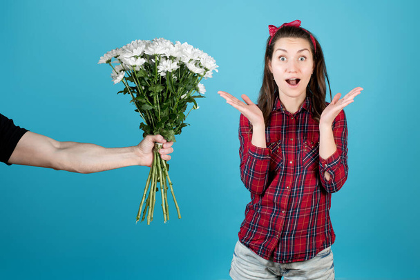 A country girl in a red plaid shirt is surprised at the gift in the form of flowers of white chrysanthemums, widening her eyes and spreading her hands. Isolated on a blue background - Photo, image