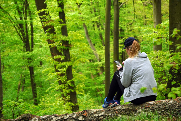 Single girl in sports uniform with cap is sitting on fallen tree, using wired headphones and looking at phone. Recreation after physical activities in nature. Green spring leaves are on background - Photo, Image