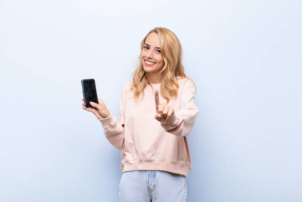 young blonde woman smiling proudly and confidently making number one pose triumphantly, feeling like a leader using a smartphone - Фото, зображення