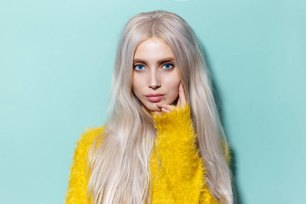Studio portret młodej blondynki z niebieskimi oczami w żółtym swetrze na tle aqua menthe kolor. - Zdjęcie, obraz