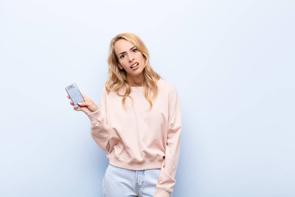 young blonde woman feeling puzzled and confused, with a dumb, stunned expression looking at something unexpected using a smartphone - Foto, Bild