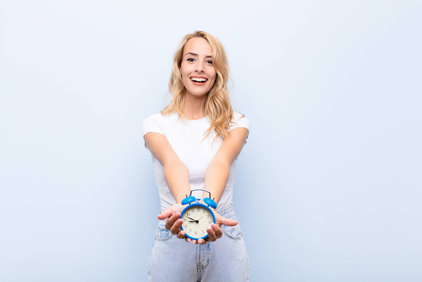 young blonde woman smiling happily with friendly, confident, positive look, offering and showing an object or concept holding a clock - Valokuva, kuva