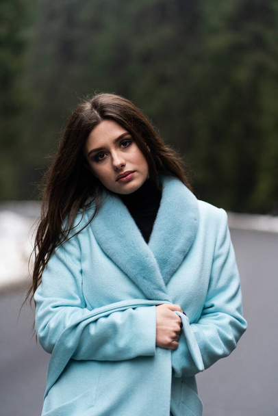 Sexy brunette girl poasing on road on winter mountains background. Glamorous young woman wearing stylish blue long coat. Fashion, business and beautiful people concept.. - Photo, Image