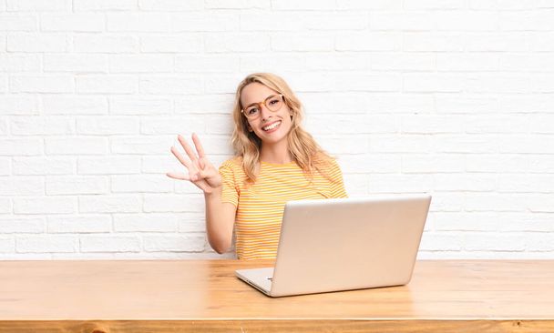 young blonde woman smiling and looking friendly, showing number four or fourth with hand forward, counting down using a laptop - Valokuva, kuva