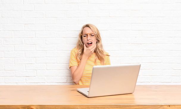 young blonde woman with mouth and eyes wide open and hand on chin, feeling unpleasantly shocked, saying what or wow using a laptop - Foto, afbeelding