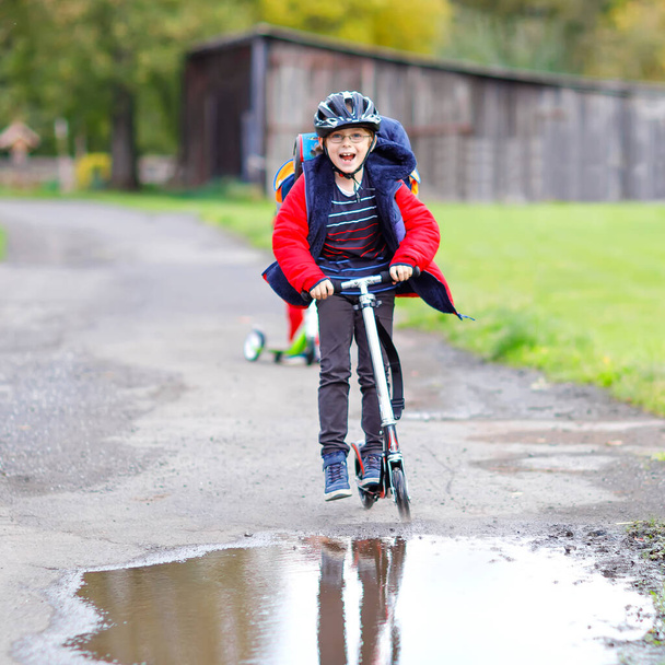 roztomilý malý školák na skútru na cestě do školy nebo ze školy. Sedmiletý školák projíždí kaluží deště. vtipné šťastné dítě v barevné módní oblečení a s helmou. - Fotografie, Obrázek
