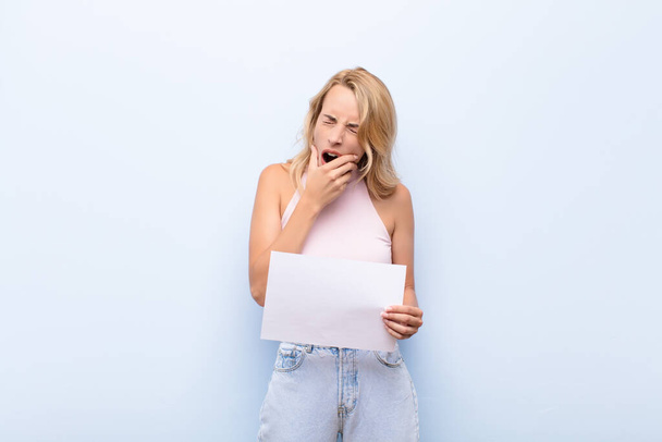 young blonde woman with mouth and eyes wide open and hand on chin, feeling unpleasantly shocked, saying what or wow holding a sheet of paper - Фото, изображение
