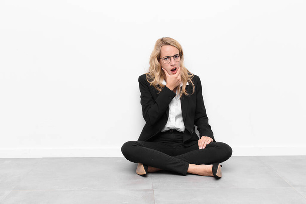 young blonde woman with mouth and eyes wide open and hand on chin, feeling unpleasantly shocked, saying what or wow sitting on the floor - Foto, Imagem