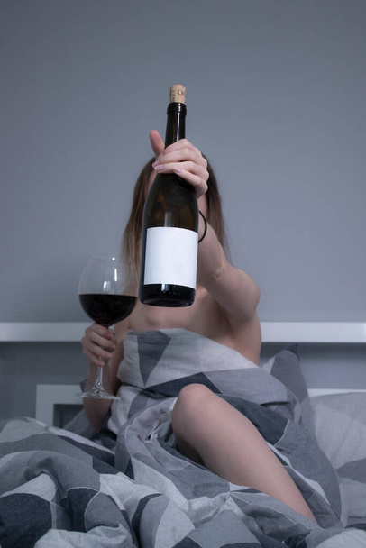 happy beautiful slender half-naked girl shows us a bottle of wine without a label, in bed with gray sheets in triangles - Photo, image