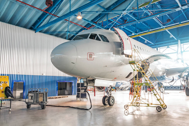 Aviones en reparación en un hangar de mantenimiento. Vista de la nariz y la cabina, puerta principal abierta con una escalera técnica
 - Foto, Imagen