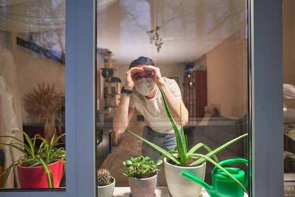 Homem com máscara médica em seus rostos olhando através da janela, auto-isolamento para evitar a propagação da infecção
. - Foto, Imagem