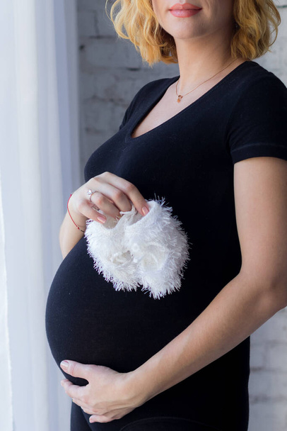 těhotná žena objímá své břicho a drží v rukou dětské ponožky. Máma čeká dítě. šťastný těhotný - Fotografie, Obrázek