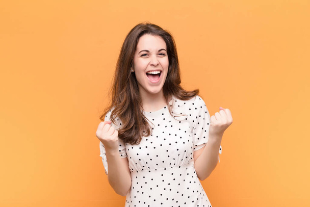 young pretty woman shouting triumphantly, laughing and feeling happy and excited while celebrating success against orange wall - Фото, изображение