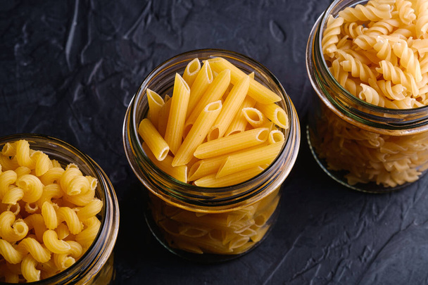 Three glass jars in row with variety of uncooked golden wheat pasta on dark black textured background, angle view macro - Fotó, kép