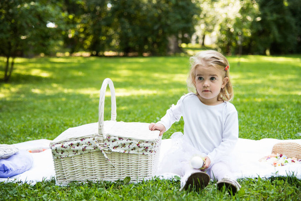 A little beautiful girl walks in the park with a picnic basket and straw hat. Funny emotional baby. - Фото, изображение