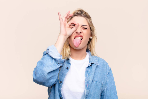 young pretty blonde woman smiling happily with funny face, joking and looking through peephole, spying on secrets against flat wall - Fotoğraf, Görsel