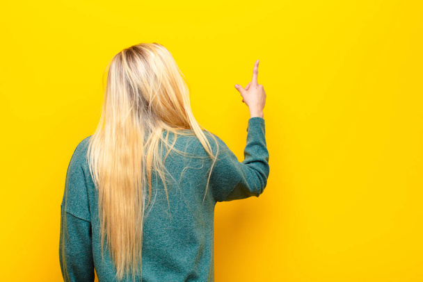 young pretty blonde woman standing and pointing to object on copy space, rear view against yellow wall - Foto, afbeelding