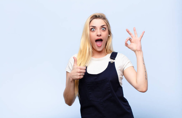 young pretty blonde woman feeling happy, amazed, satisfied and surprised, showing okay and thumbs up gestures, smiling against flat wall - Fotoğraf, Görsel