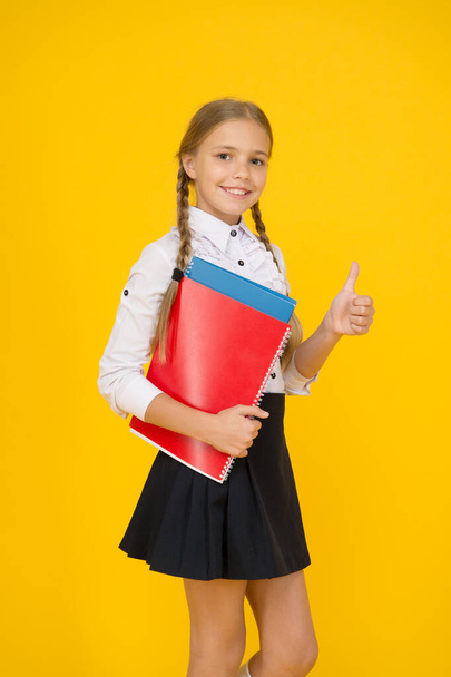 back to school. small girl in uniform show thumb up. child after lessons carry documents. study at home. school fashion style. smart looking student yellow background. modern education concept - Photo, image