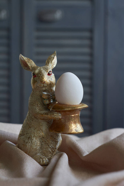Frühlingskomposition, goldene Hasenfigur mit Ei auf beiger Tischdecke, Osterkonzept, selektiver Fokus - Foto, Bild