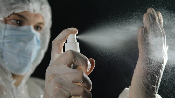 Médico masculino con una máscara médica y traje protector trata sus manos con un antiséptico. pandemia de Coronavirus
. - Foto, Imagen