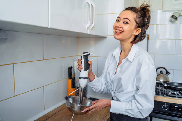 Attractive girl preparing dough in a food processor while standing in a bright kitchen. Mixing dough with hand mixer on kitchen table background with cooking ingredients - Foto, Imagen