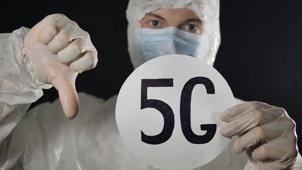 Man in a medical mask and protective suit raises the 5 g sign and shows his thumb down. 5 g networks spread the coronavirus. - Photo, Image