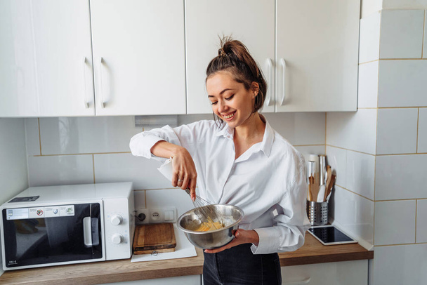 Красивая, привлекательная, улыбающаяся молодая женщина смешивает тесто с венчиком для банановых блинов в стальных блюдах на кухне с белым, светлым интерьером. Монохромное фото без ярких акцентов
 - Фото, изображение
