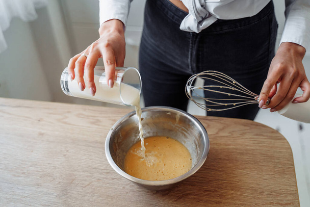 Concept de cuisine maison bricolage. Une femme brune lumineuse tamise la farine pour les crêpes à travers un tamis, salant la pâte, ajoutant du lait. Jeune femme souriante, aime cuisiner dans une belle cuisine moderne
. - Photo, image