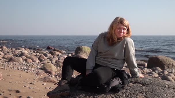 Young girl woman with blond hair sitting near sea and grimaces. warm shirt - Footage, Video
