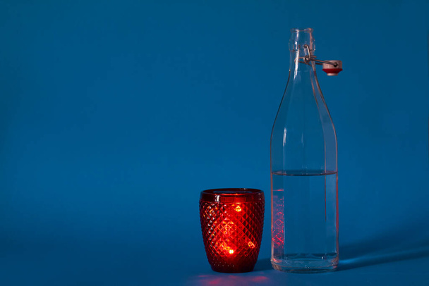 Lasipullo, jossa kirkas vesi ja punainen lasi sinisellä taustalla, valikoiva painopiste
 - Valokuva, kuva