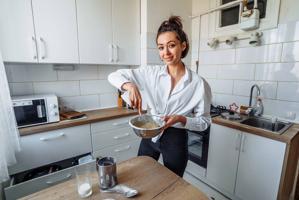 Ελκυστική κοπέλα ετοιμάζεται ζύμη σε ένα επεξεργαστή τροφίμων, ενώ στέκεται σε μια φωτεινή κουζίνα. Ανάμιξη ζύμης με μίξερ χειρός σε φόντο τραπεζιού κουζίνας με υλικά μαγειρέματος - Φωτογραφία, εικόνα