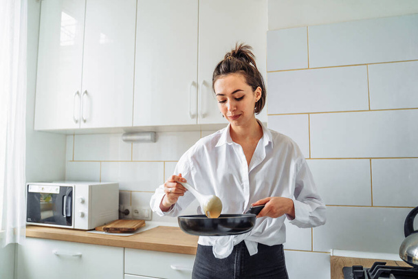 Woman pours the dough into a pan to cook homemade fried banana pancakes. Hobby in a beautiful bright kitchen. The economic wife is waiting for her husband from work with delicious tasty food - Photo, Image
