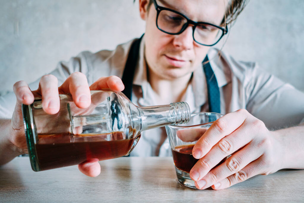 Egy szemüveges férfi whiskyt vagy konyakot önt egy pohárba egy kemény munkanap után. Közelről.. - Fotó, kép