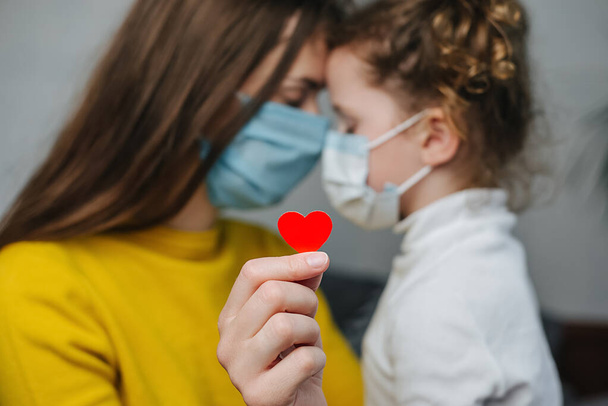 Young mommy hugs cute preschool daughter in protective medical masks sitting on bed, holding red heart a way to show appreciation and to thank all essential employees during Covid-19 pandemics - Foto, imagen