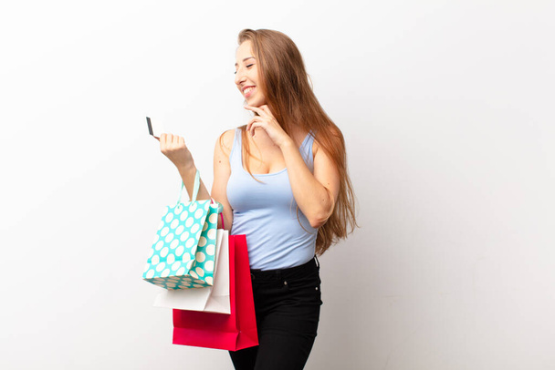 yound blonde woman smiling with a happy, confident expression with hand on chin, wondering and looking to the side holding shopping bags - Foto, Imagem