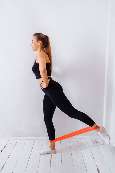 The concept of sports training at home. The girl does exercises for the arms and legs using fitness gum. Strengthens the muscles in the body with a red color rubber band with strong resistance - Foto, Bild