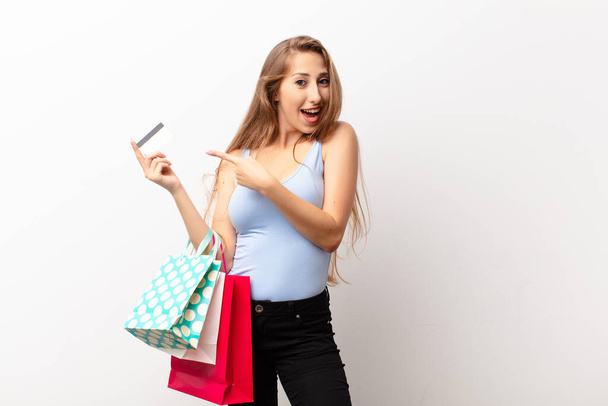 yound blonde woman looking excited and surprised pointing to the side and upwards to copy space holding shopping bags - Фото, зображення
