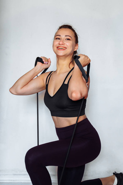 Muscular, attractive, beautiful blonde fitness trainer doing home exercises with a black expander. She crouching and kicking up, smiling. Woman exerting a lot of strength to make her body perfect. - Photo, Image